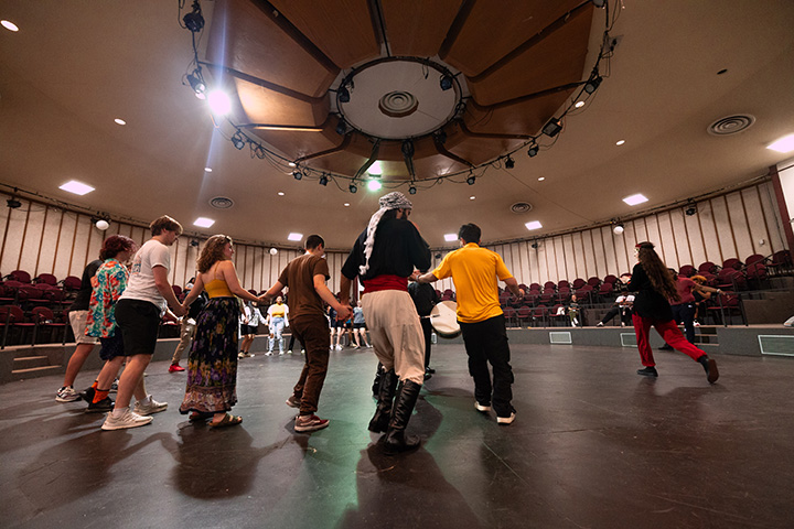 A group of a dozen students stand on a large stage learning a Middle Eastern dance.