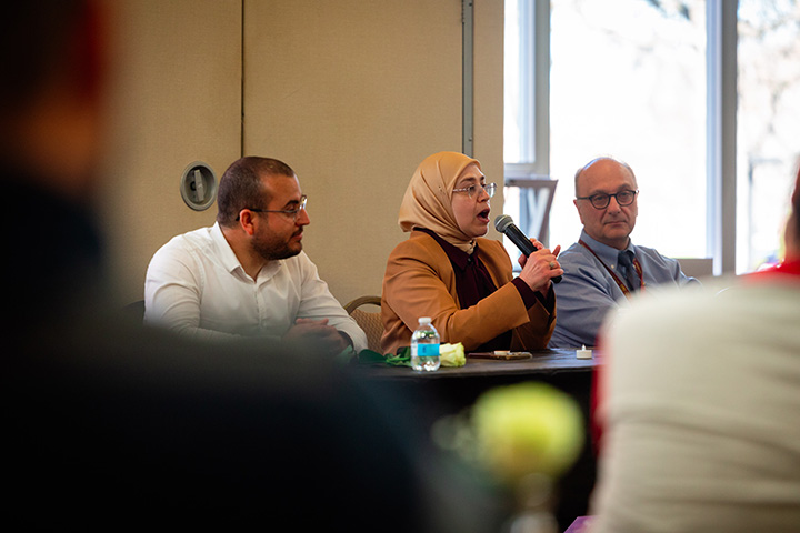 Three people sit at a table speaking to a group as a woman holds a microphone.