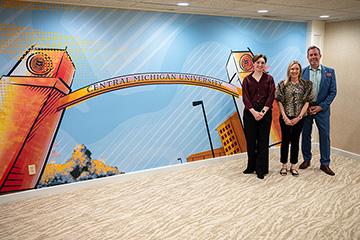 The CMU president, first lady and an art student stand in front of a CMU-themed mural painted on a wall.