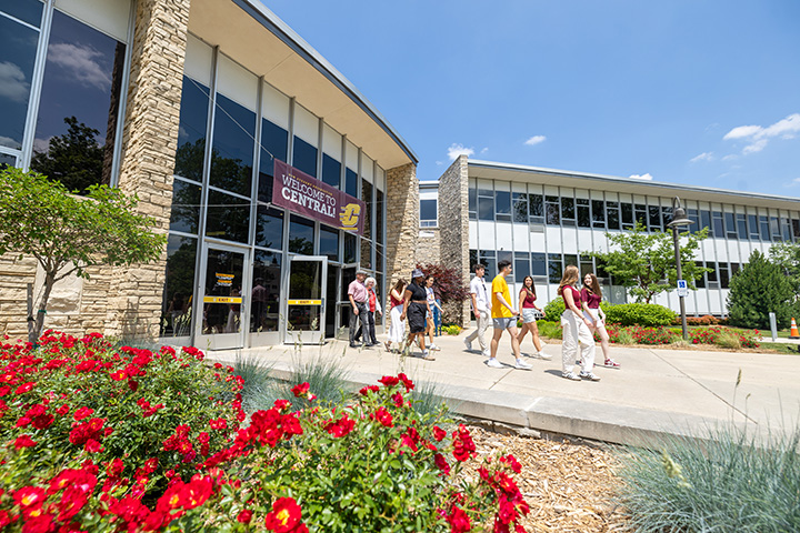 A group of students and parents walk out of the University Center. Trees and flowers are in bloom along the walkway.