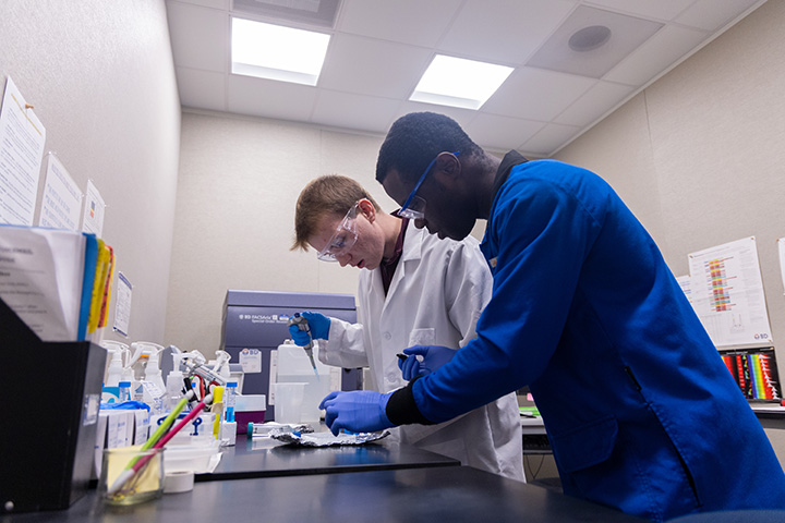 Two people in lab coats and goggles inside a lab.