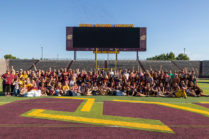 A large group of CMU alumni pose for a photo inside the football stadium with a large scoreboard in the background.