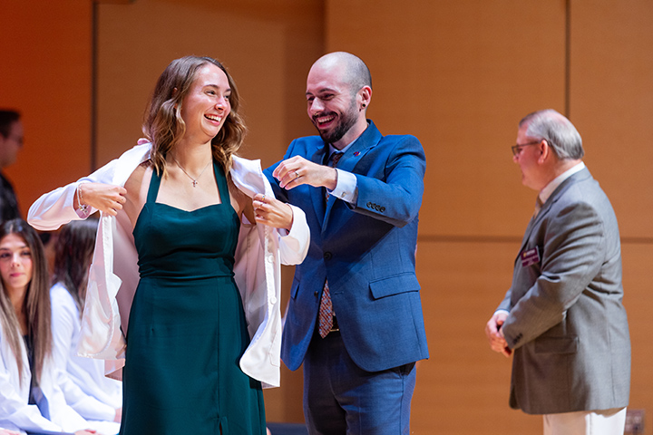 A man in a suit helps woman in a dress put on her white coat.