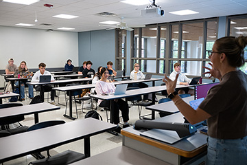 A group of people sitting in a classroom at tables, looking at a professor talk.