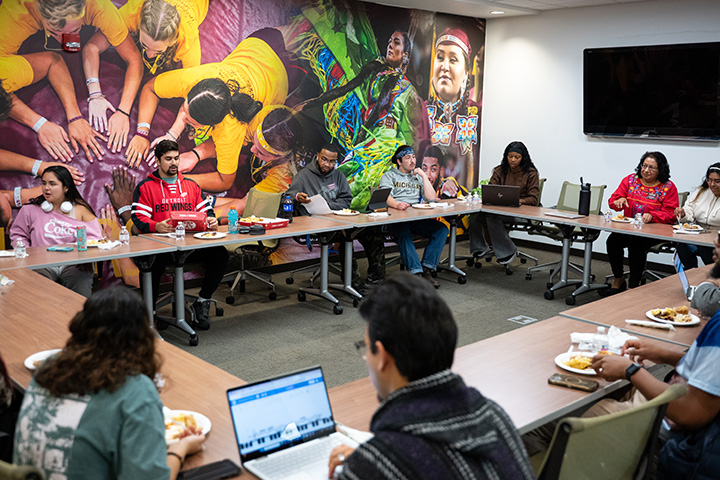 A large group of people sit at rectangular tables set up in a circle inside a room.