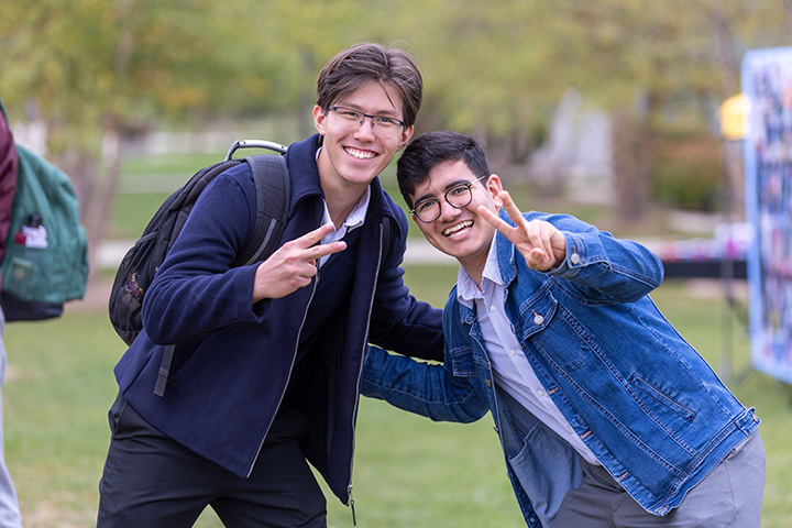 Two male college students wearing jackets and glasses smile for the camera flashing peace signs while standing outside