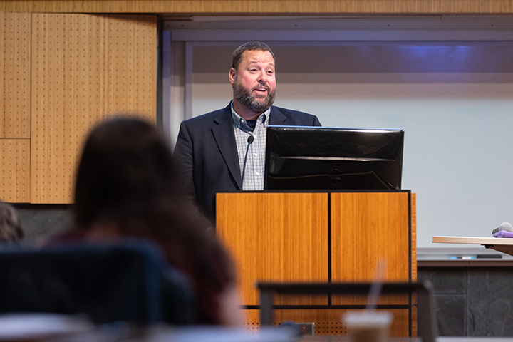 A man in a sport coat and collared shirt stands at a podium addressing people in an auditorium