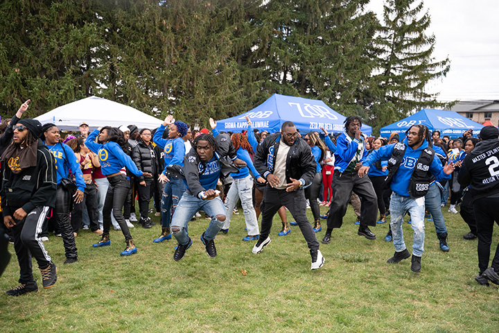 A group of people dancing and jumping around on the grass in front of some large trees