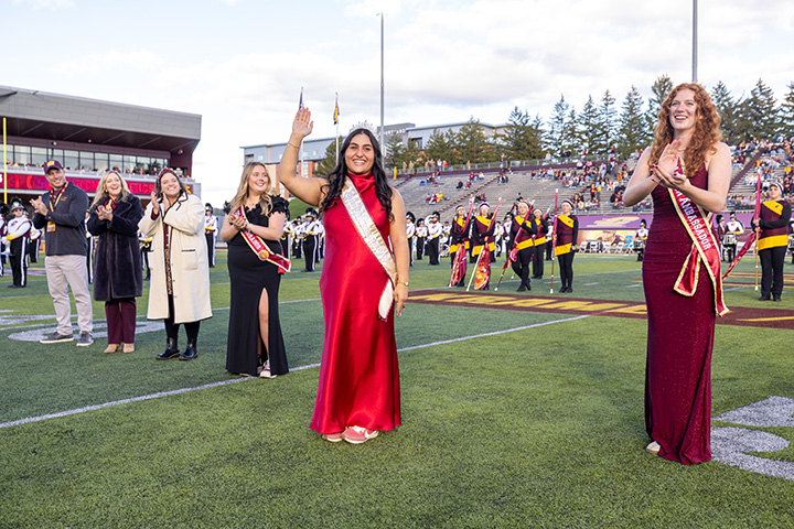 A group of people, including a woman in a red dress stand on a football field waving to the crowd