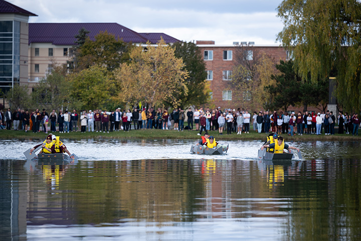 Three pairs of people sit in small cardboard boats paddling on a pond.