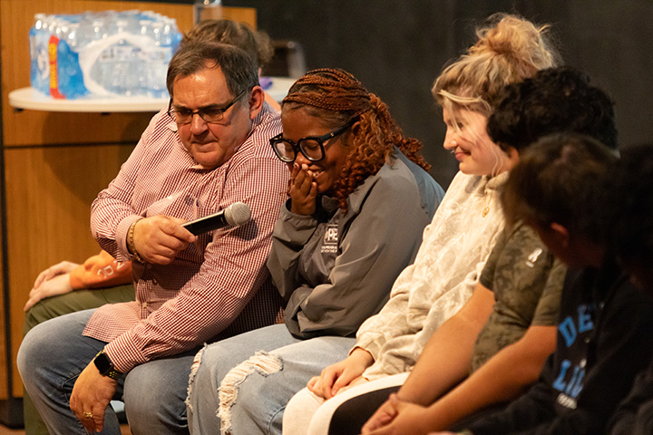A man in a red and white striped shirt sits in a chair pointing a microphone at a young woman who is laughing. Three other people sit nearby.