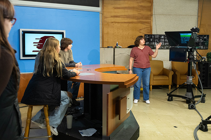 Agroup of people in a a television studio. Two sit at a desk. Another stands near a TV camera.