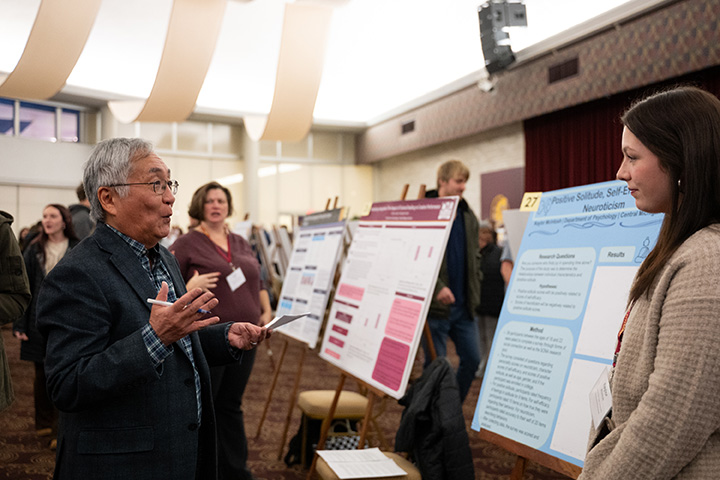 A man and female student standing in front of a display board inside a large room.