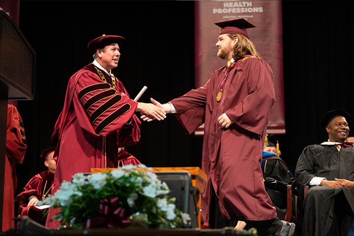 Two men shaking hands wearing maroon graduation gowns.