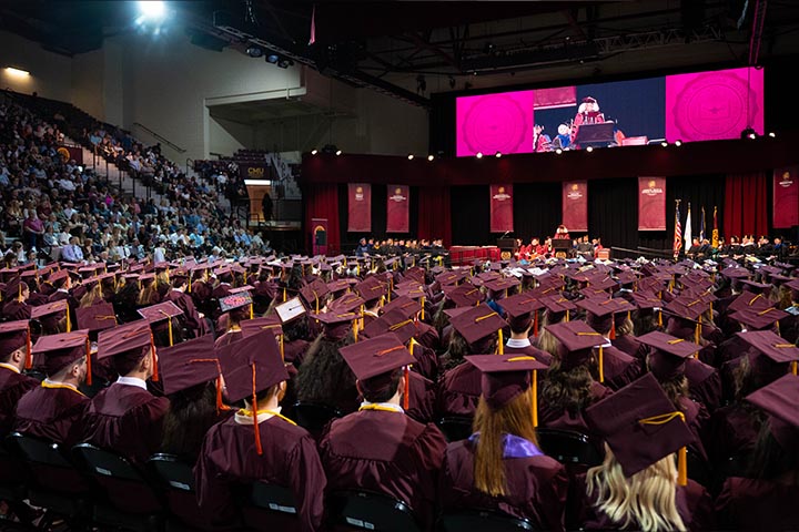 A large group of people in maroon graduation gowns and caps facing a large video screen..