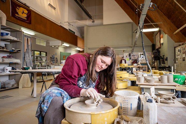 A pottery student sits at a pottery wheel making a clay bowl.