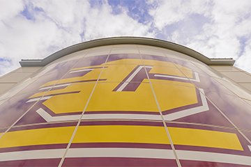 A tall building with a maroon and gold logo on it as clouds pass overhead.