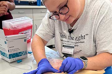 A woman wearing blue nitrile gloves places a needle in a plastic arm