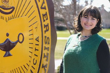 A woman in a green sweater smiling in front of a university sign in the sunshine.