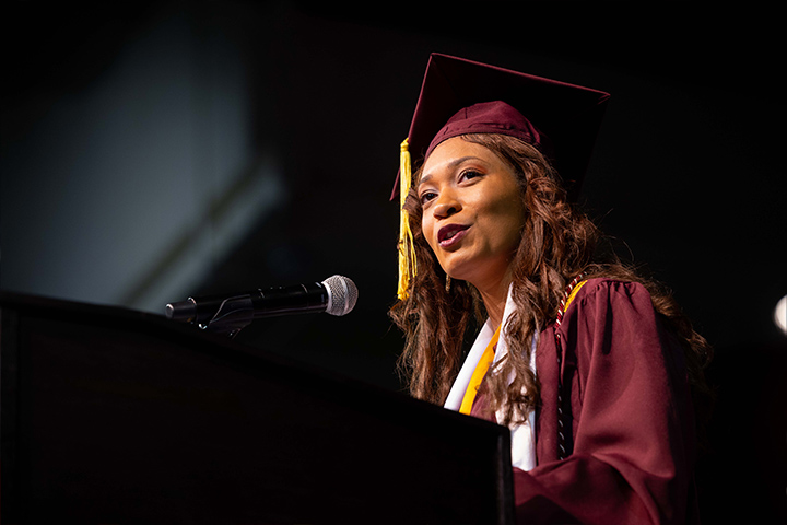 A woman in a graduation gown and cap speaking into a microphone.