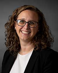 A smiling woman with blond curly hair wearing a white shirt and black blazer