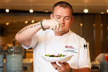 A chef in a white uniform sprinkles seasoning onto a salad