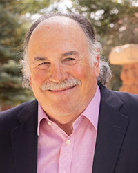 A close-up photo of a smiling man wearing a pink shirt and blue coat