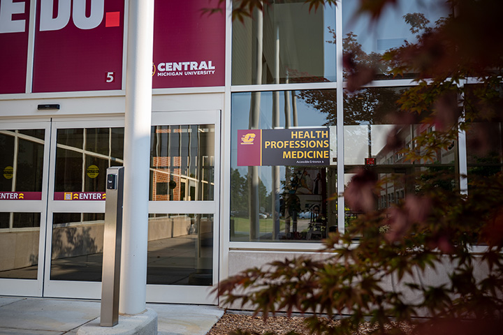 The front entrance of a building with glass windows and doors and a sign that reads Health Professions Medicine.