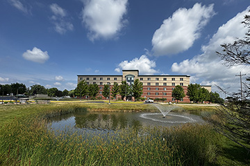A brick college residence hall building with a pond and fountain in the foreground. Blue skies and big white clouds are overhead.