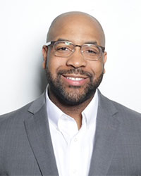 A close-up image of a smiling man wearing glasses and a grey suit