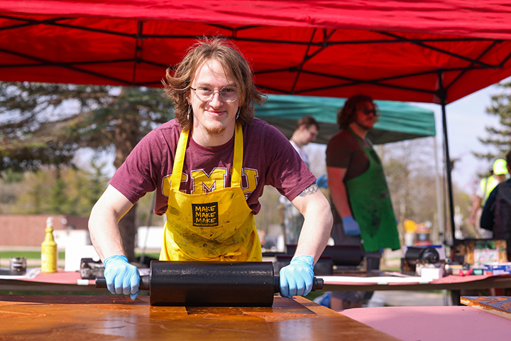 A CMU student wearing a CMU t-shirt and yellow apron holds a large rolling pin while pressing art prints.