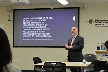 A man in a suit presenting information in front of a large screen.