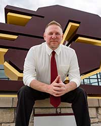 A man wearing a white shirt, red tie and black pants sits in front of a large maroon-colored letter C