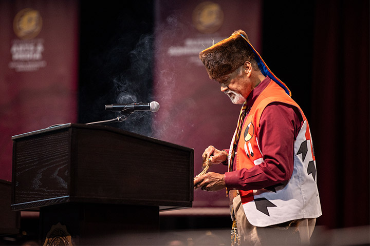 A man wearing a red and white vest standing at a podium and burning sage.