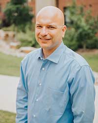 A bald man in a blue button-up shirt smiles into the camera