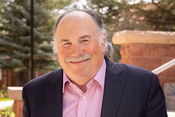 A close-up photo of a smiling man wearing a pink shirt and blue coat