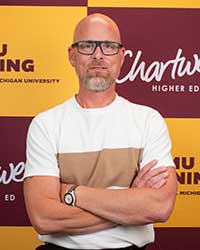 A man wearing glasses and a white shirt stands with his arms crossed in front of a maroon and gold backdrop