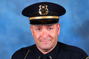 A man in a police uniform smiles at the camera in front of a blue background.