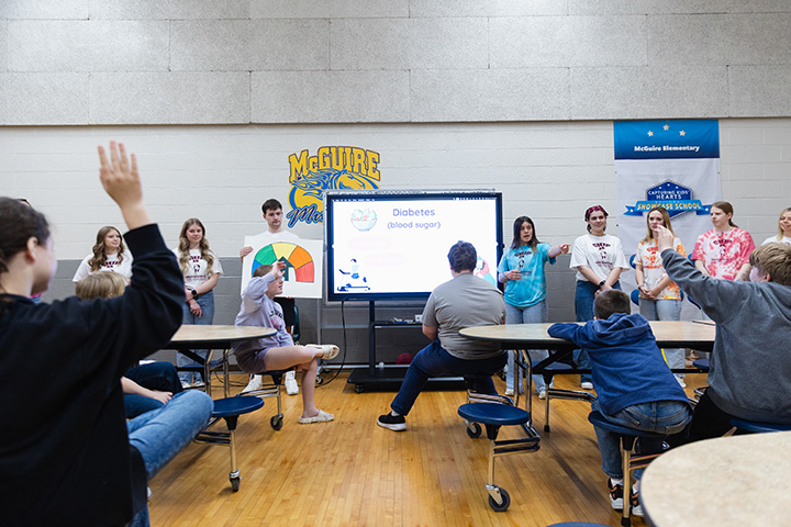 CMU Exercise is Medicine students stand in front of a group of elementary students inside a lunchroom