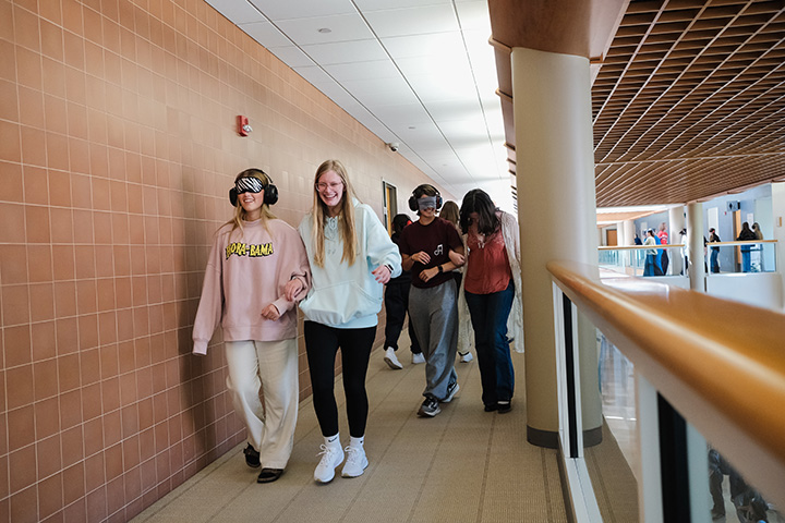 Pairs of students walk down a hallway, one wearing a blindfold and earmuffs, the other leading the way