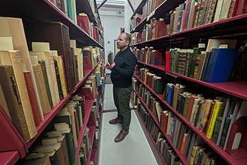 A man in a dark shirt and dark pants stands in the middle of two six-shelf units full of books.