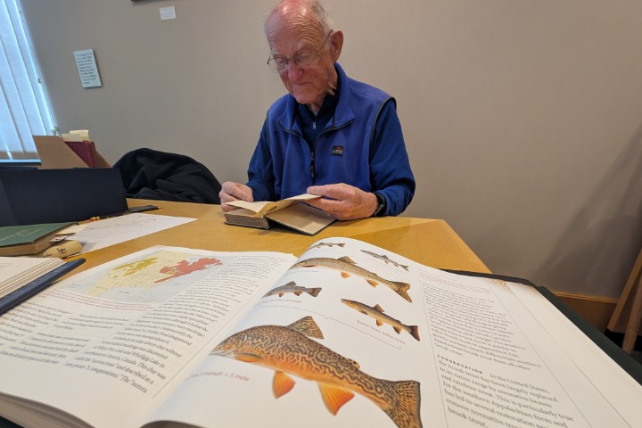 A man in glasses reads an open book at a table with another book with pictures of fish point toward the camera.