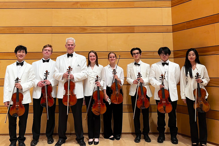 Seven people wearing black bowties and pants and white jackets and shirts, and holding violins, pose in front of a two-tone brown wall.