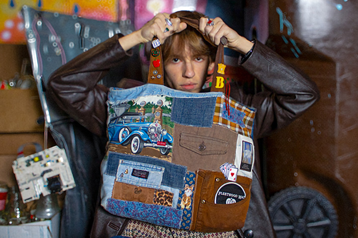 A young man in a brown leather jacket holds a patchwork cloth bag in front of him with both hands.