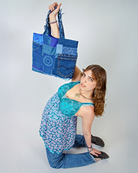 A young woman dressed in blue kneels on the ground while holding a patchwork denim bag with her right hand.