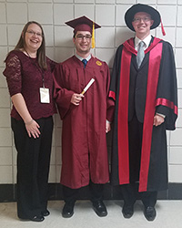 A young man in glasses wearing a maroon graduation cap-and-gown ensemble stands next to a woman in a maroon top and black pants and long hair with a man in a black-and-maroon academic robe on his other side.