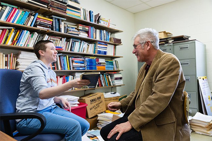 A young woman in a gray sweatshirt and blue jeans sitting in a blue office chair hands a book to a seated gray-haired man with glasses and a brown blazer.