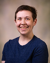 A woman with short-cropped hair and a blue top smiles against a brown background.