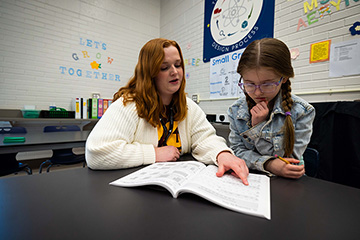 A woman with red hair and a white sweater points to something in an open book sitting on a table while a young girl with glasses and pig tails looks on.