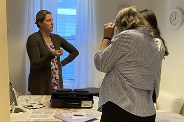 A woman in a three-quarters sweater over a floral top stands behind a table and talks to two other women.